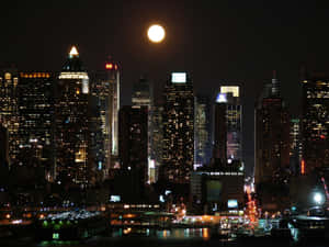 滿月照耀在夜晚燈火通明的城市天際線上，下方有水。 — 高譚 壁紙