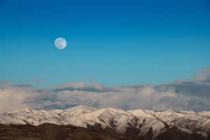 滿月高掛在積雪的山脈和雲層之上，天空清澈湛藍。 — 月光 壁紙