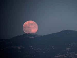 粉紅色滿月從黑暗的山丘上升起，下方是城鎮。 — 月亮與星星 壁紙