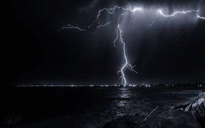 閃電照亮夜間暴風雨的海洋，遠處有城市燈光。 — 閃電麥坤 壁紙