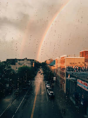 彩虹橫跨濕漉漉的城市街道，透過雨滴窗戶觀看 — 彩虹美學 壁紙