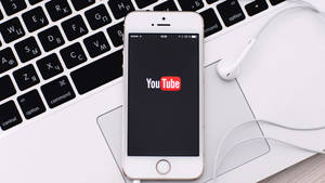 白色手機螢幕上顯示YouTube應用程式，放在筆記型電腦鍵盤上，耳機在附近。 — YouTube 標誌 壁紙