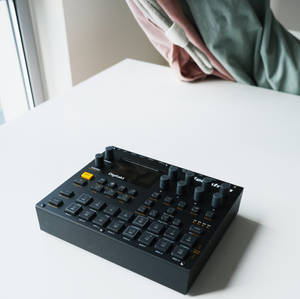 窗邊白色桌上的黑色 Digitakt 音樂序列器，帶有垂掛布料。 — 低傳真 壁紙