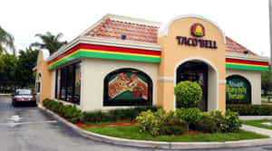 Taco Bell餐廳，米色牆壁和紅色瓦片屋頂，周圍環繞著綠色植物。 — 塔可 壁紙