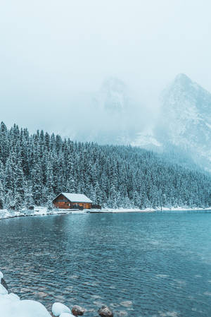冬季藍色湖邊的雪中小屋，有松樹和霧山。 — 冬季風景 壁紙