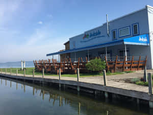 一間海濱餐廳，在平靜水邊的碼頭上有木製桌椅。 — 湖泊 壁紙
