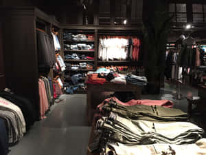 一間時尚服飾店，在溫暖燈光下展示著掛架和成堆的衣物。 — Hollister 壁紙