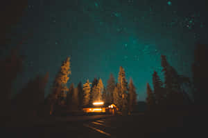 一間燈光溫暖的小屋，被松樹環繞，上方是星空。 — 夜空 壁紙