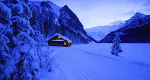 暮色中雪山山谷裡的雪覆蓋小屋，溫暖燈光閃爍。 — 冬季風景 壁紙