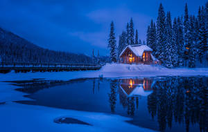 暮色中，溫暖燈光映照在結冰湖面上的雪中小屋。 — 冬季風景 壁紙