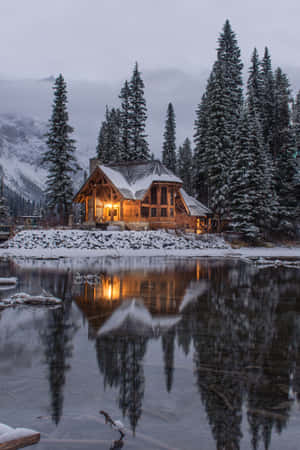 舒適的小屋，溫暖的燈光倒映在冰湖上，四周環繞著白雪覆蓋的松樹。 — 暮光 壁紙
