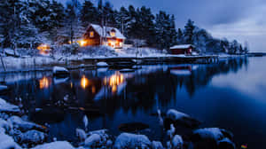 白雪覆蓋的湖邊小屋，溫暖的燈光在夜晚平靜的水面上反射。 — 冬季美學 壁紙