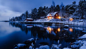 雪覆蓋的湖邊小屋，溫暖燈光在夜晚平靜的水面上反射。 — 冬季風景 壁紙