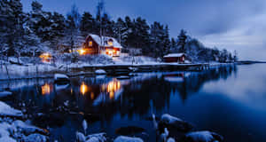 夜間水邊的雪地小屋，溫暖燈光倒映在平靜水面 — 冬季風景 壁紙