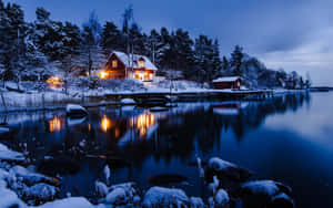 夜間湖邊被雪覆蓋的小屋，溫暖燈光倒映在水中 — 冬季美學 壁紙