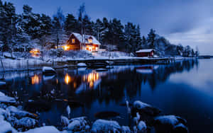 夜晚湖邊雪覆蓋的小屋，溫暖燈光反射在平靜水面上。 — 冬季房屋 壁紙