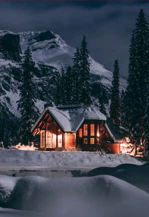 夜晚，一間溫暖照明的木屋坐落在白雪覆蓋的山林之中。 — 冬季風景 壁紙