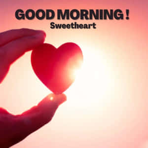 手拿著一個帶有'GOOD MORNING Sweetheart'文字的紅色心形，背景是日出。 — 早安我的愛 壁紙