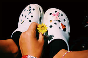 手在裝飾過的白色Crocs附近拿著一朵黃花。 — Crocs 壁紙