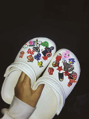白色Crocs涼鞋裝飾著卡通和超級英雄貼紙，由一隻手拿著。 — iPad Pro M4 壁紙