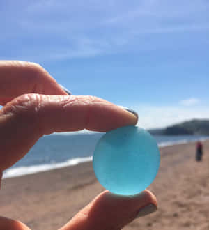 手在陽光明媚的海灘上拿著藍色海玻璃，背景有海洋和山丘。 — 海灘 壁紙