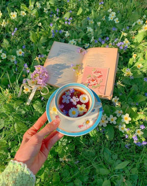 手拿著花卉茶杯，旁邊放著打開的書，在長滿野花的草地上。 — 花 iPhone 壁紙