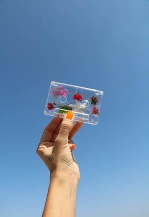 手拿著一個透明的卡式錄音帶，裡面有花，背景是藍天。 — OnePlus Pad 4 壁紙