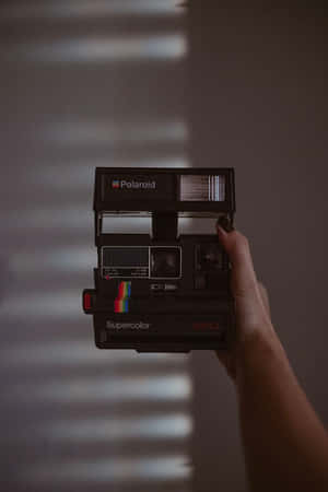 手拿著黑色Polaroid Supercolor相機，後面牆上有光條。 — 拍立得 壁紙
