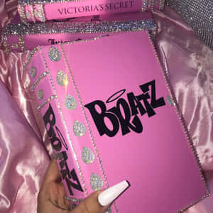手拿著一本帶有 Victoria's Secret 品牌標誌的粉紅色閃亮 Bratz 書，放在緞布上。 — 復古90年代風格 壁紙