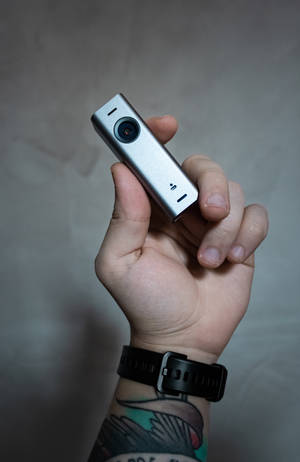 手拿銀色運動相機，有刺青手腕和黑色手錶。 — 榮耀MagicPad 3 Pro 12.3 壁紙