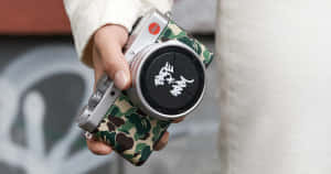 手拿著帶有迷彩機殼和塗鴉鏡頭蓋的銀色 Leica 相機。 — Bape 動態 壁紙