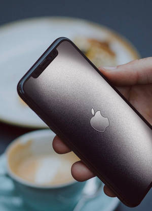 手在咖啡和糕點附近拿著深灰色iPhone。 — 蘋果標誌 iPhone 壁紙