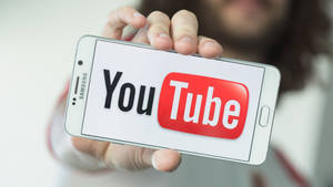 手拿著顯示YouTube應用程式標誌的智慧型手機。 — 社群 壁紙