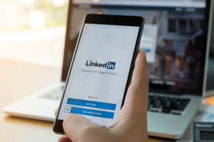 手拿著電話顯示LinkedIn登入畫面，背景有筆記型電腦。 — 領英 壁紙