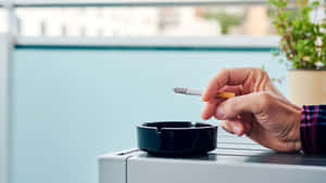 手拿著點燃的香煙在黑色煙灰缸上，陽台有城市景觀和植物。 — 抽菸 壁紙