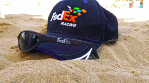 在明亮陽光下，沙灘上的FedEx Racing帽和太陽眼鏡。 — 聯邦快遞 壁紙