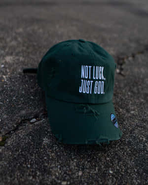 一頂綠色破舊的帽子，上面有白色文字寫著 not luck just god，在破裂的瀝青路面上。 — Apple Watch Ultra 壁紙