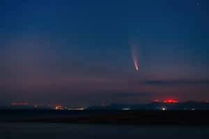 一顆彗星在黑暗海岸線上的夜空中發光，遠處有城市燈光。 — MacBook Air M3 壁紙