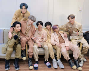 七位穿著米色服裝的K-pop偶像在沙發上擺姿勢，穿著各種鞋子。 — 5:4 壁紙
