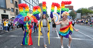 三位變裝表演者穿著彩虹服裝，手持拼出PRIDE的氣球字母遊行。 — 慶祝 壁紙