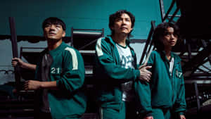 三位身穿綠色號碼運動服的人在黑暗的工業環境中手持武器。 — 067 魷魚遊戲 壁紙