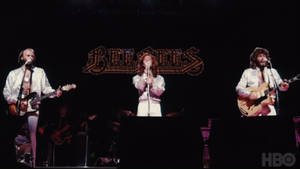 Bee Gees 身穿白色服裝，手持吉他和麥克風在舞台上現場表演。 — 好萊塢 壁紙