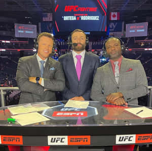 三位身穿西裝的男性UFC評論員在體育場館的ESPN轉播桌旁。 — 運動 壁紙