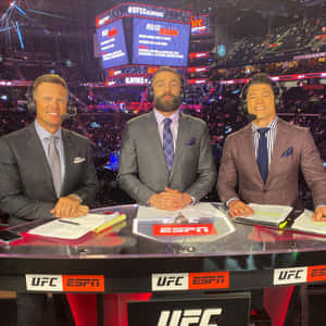 三位身穿西裝的男性UFC評論員在燈光明亮的競技場轉播桌前。 — 終極格鬥冠軍賽 壁紙