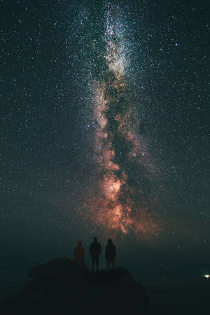 三個人以剪影形式出現在山頂上，下方是夜晚充滿活力的銀河。 — 三星Galaxy 壁紙