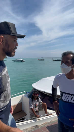 三個男人在熱帶碼頭的船上，周圍有藍綠色海水和遠處的船。 — 運動 壁紙