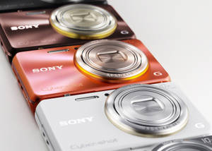 三款Sony Cyber-shot相機，玫瑰金、橙色和白色，在白色背景上。 — Sony 壁紙