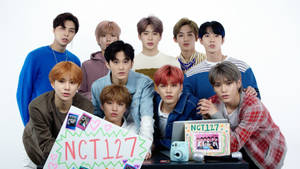 九位K-pop偶像在攝影棚內手持NCT127標語牌和相機擺姿勢。 — Nct 127 壁紙