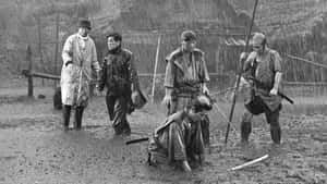 五個身穿傳統武士裝備的男人在暴雨中站立和跪在泥濘的田野上。 — 黒死牟 壁紙
