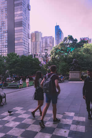 日落時分，人們在棋盤格廣場上行走，背景是城市建築和樹木。 — 暗紫色美學 壁紙